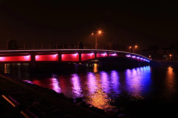 Illuminated mystical bridge at night with vibrant purple and red lights reflecting over calm water.