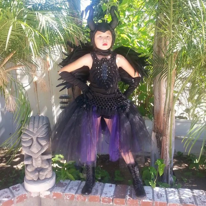 Child in detailed black and purple Halloween costume with horns and wings posing outdoors among green plants.