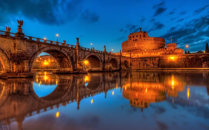 Historic mystical bridge and ancient castle illuminated at dusk, reflecting beautifully on calm river waters.