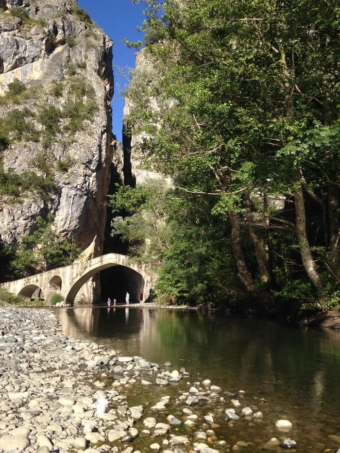 Ancient mystical stone bridge over calm river between rocky cliffs and lush green trees on a sunny day.