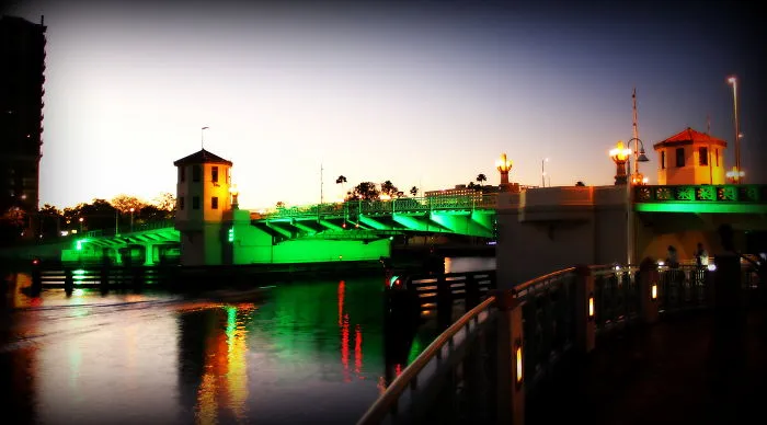 Mystical bridge illuminated with green lights at twilight, reflecting on calm water, creating a magical atmosphere.