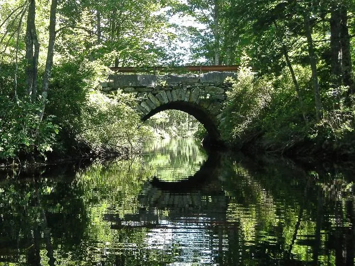 Stone arch mystical bridge over calm water surrounded by lush green trees reflecting another world.