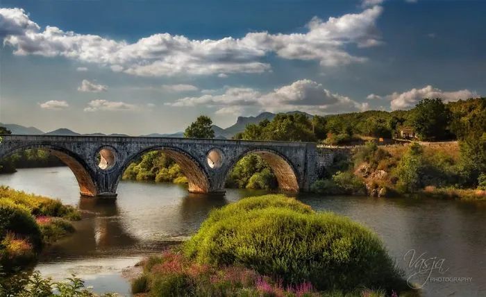 Ancient stone mystical bridge over calm river surrounded by lush greenery and dramatic cloudy sky.
