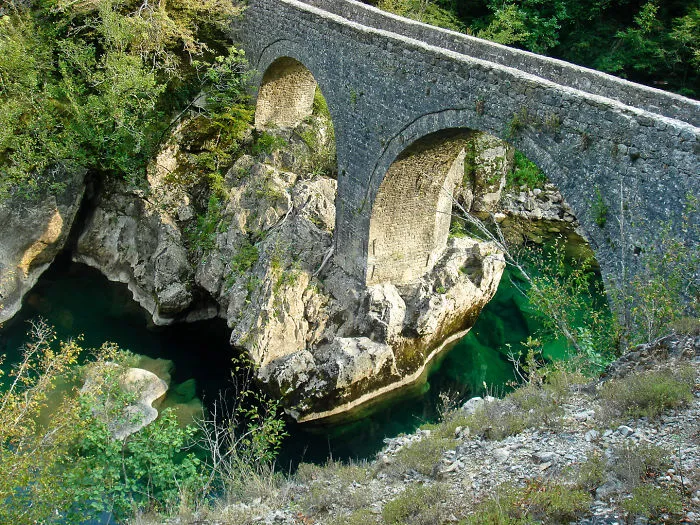 Stone arch mystical bridge over clear green river surrounded by rocks and lush forest landscape view.