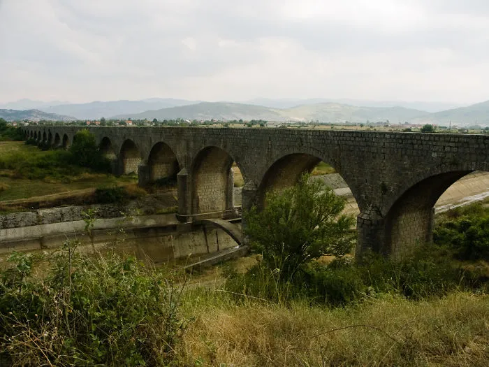 Ancient mystical stone bridge with multiple arches spanning a dry riverbed, surrounded by greenery and distant mountains.
