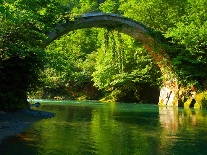 Stone mystical bridge arching over clear river surrounded by lush green trees in a serene natural setting