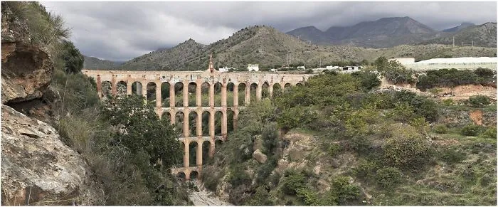 Ancient mystical stone bridge spanning a deep gorge surrounded by lush greenery and rugged mountains under cloudy skies.