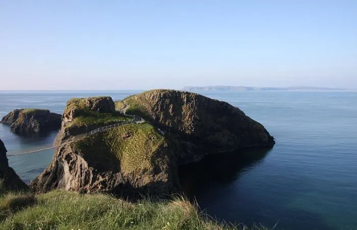 Mystical bridge connecting rocky cliffs over calm ocean waters, surrounded by green grass and clear blue sky.
