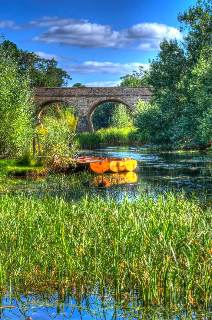Stone bridge with arches over a calm river surrounded by lush greenery and a wooden boat reflecting on the water.