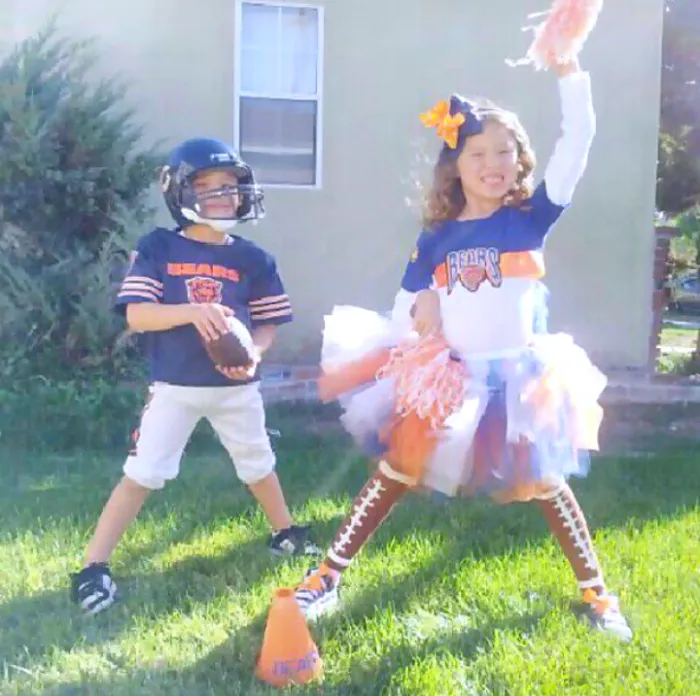 Two children dressed in Halloween costume ideas as a football player and cheerleader playing outside on green grass.