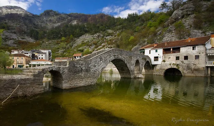 Ancient mystical stone bridge over calm river waters with mountains and village houses in the background on a sunny day.