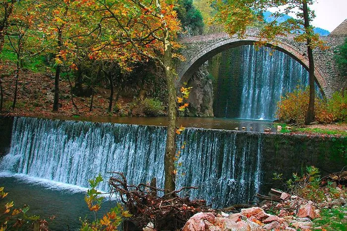 Stone mystical bridge arching over a waterfall surrounded by autumn trees and lush greenery in a serene natural setting.