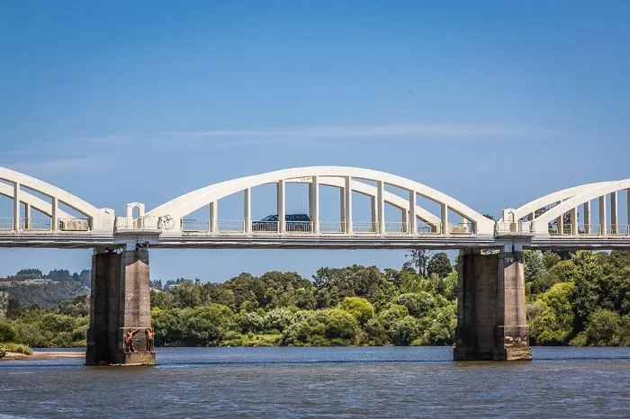 White arched bridge over calm river with green trees in the background under a clear blue sky, mystical bridge scene.