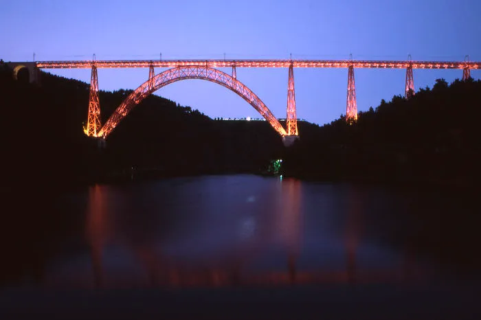 Illuminated mystical bridge arching over calm water at dusk, surrounded by dark forested hills.