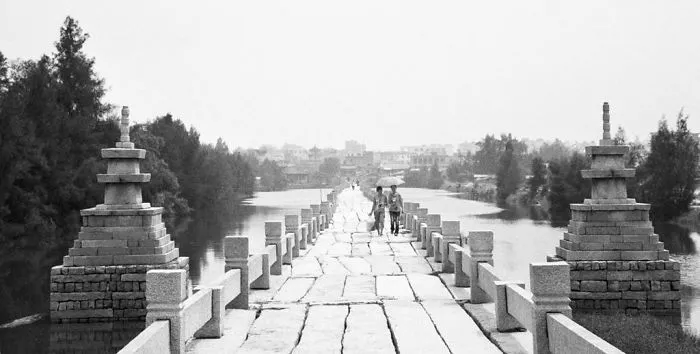 Ancient mystical bridge over calm river with two people walking, surrounded by trees and distant cityscape in monochrome.