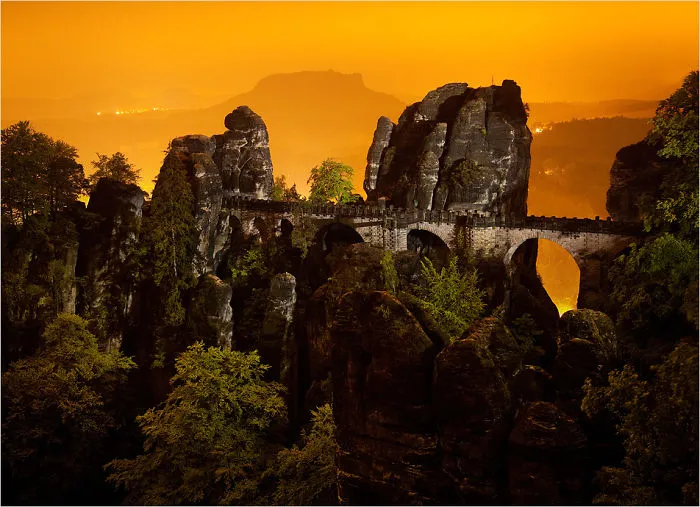 Mystical stone bridge arches over rocky cliffs surrounded by dense forest under a glowing orange sky at dusk.