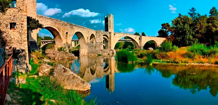 Ancient mystical stone bridge with arches reflecting over a calm river surrounded by greenery under a blue sky.