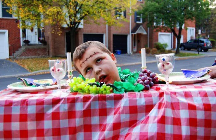 Child dressed in a Halloween costume as a spooky head on a table with grapes and candles outdoors.