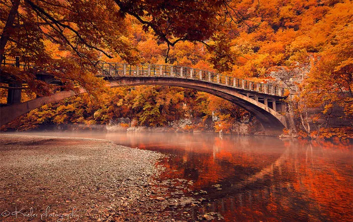 Mystical stone bridge over calm river surrounded by vibrant autumn trees and colorful foliage in a serene forest setting