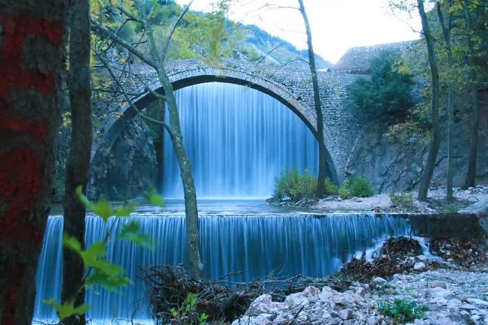 Stone arch mystical bridge over cascading waterfalls surrounded by trees and rocky terrain in a serene forest setting