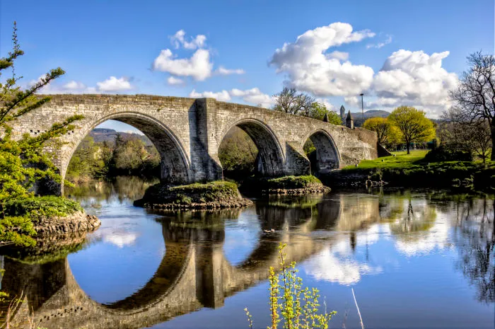 Stone arch mystical bridge over calm river surrounded by green trees under a bright blue sky with clouds.