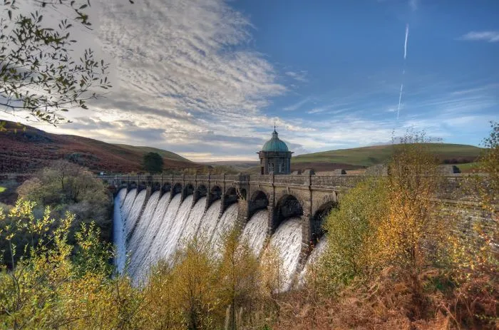 Stone arched mystical bridge with waterfall flowing underneath surrounded by autumn trees and hills under a bright sky.