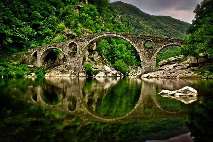 Ancient mystical stone bridge arching over calm water surrounded by lush green forest and rocky hills.