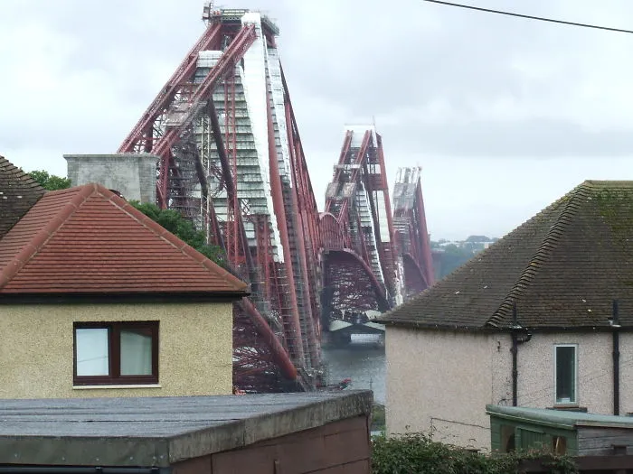Iron red mystical bridge spanning a river, framed by residential rooftops under a cloudy sky.