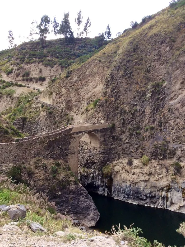 Ancient mystical stone bridge arching over dark river nestled between rugged cliffs and grassy hillsides.