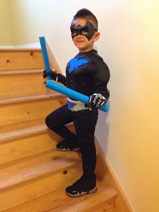 Young boy posing on wooden stairs in a black and blue superhero costume holding blue foam weapons for Halloween costume ideas.