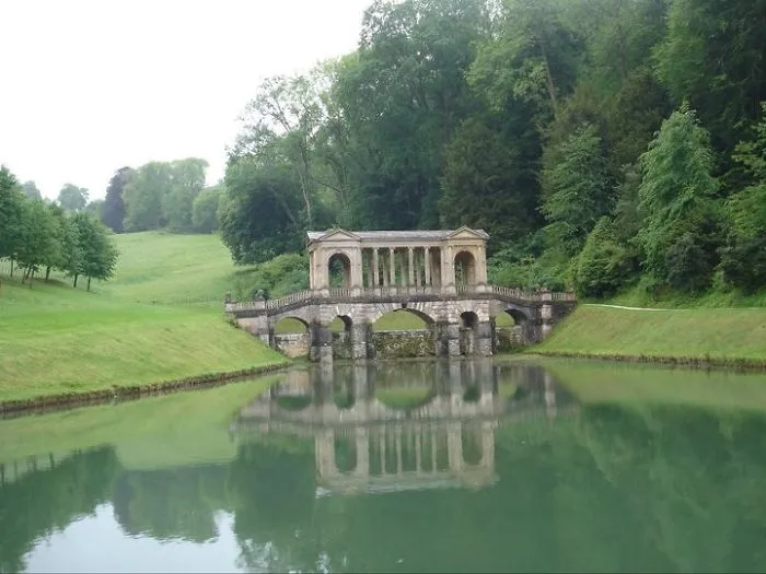 Mystical stone bridge with arches and columns reflecting on calm water, surrounded by lush green trees and grass.
