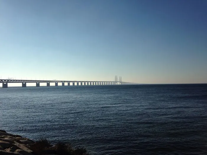 Long mystical bridge extending over calm water under a clear blue sky, creating a surreal and otherworldly scene.