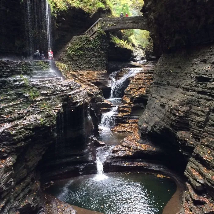 Stone mystical bridge over a cascading waterfall in a lush canyon surrounded by layered rock formations and greenery.