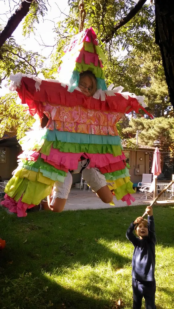 Child in colorful pinata costume hanging from tree with another child holding stick in backyard setting, children's Halloween costume ideas.