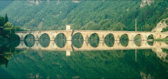 Ancient mystical bridge with multiple arches reflected in calm water surrounded by lush green hills.