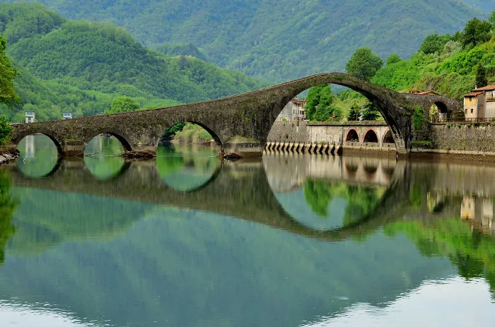 Ancient mystical bridge with stone arches over calm river, surrounded by lush green mountains and village reflections.
