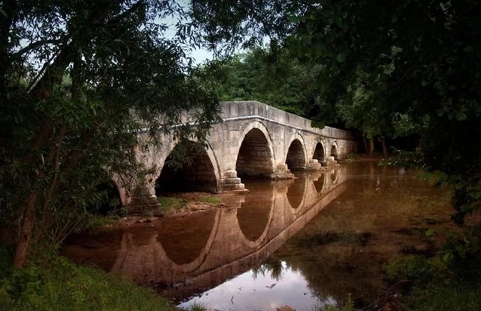Ancient stone mystical bridge with multiple arches reflecting in calm water surrounded by lush green forest landscape.