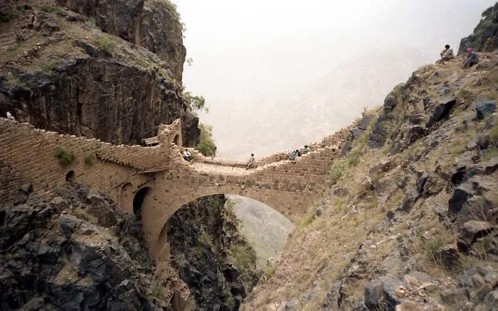 Ancient mystical stone bridge spanning a deep rocky gorge with people sitting and exploring the scenic natural landscape.