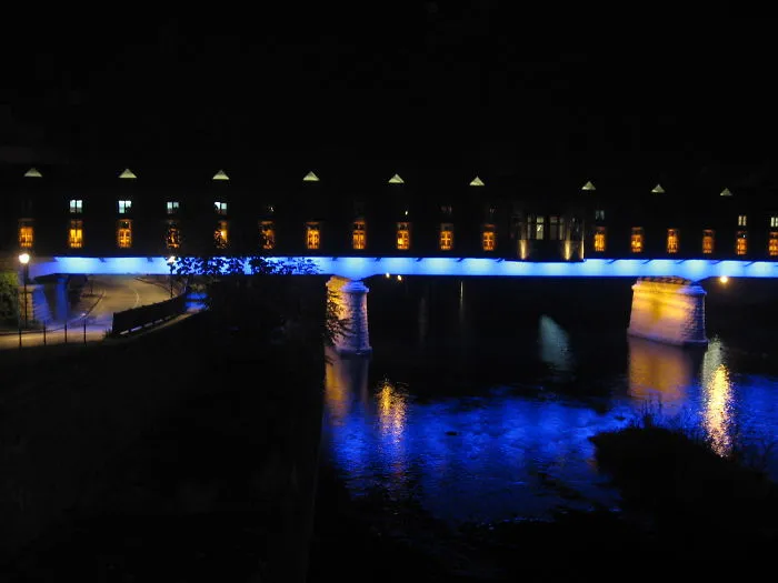 Mystical bridge glowing blue over dark river at night with illuminated building windows in the background.