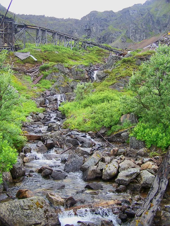 Rustic mystical bridge over a rocky mountain stream surrounded by lush greenery and rugged terrain.