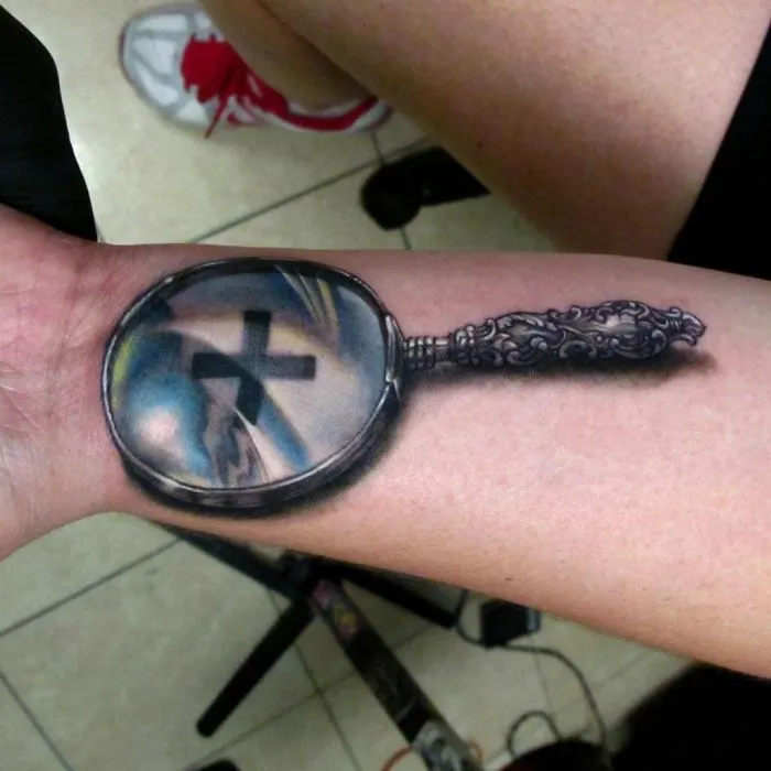 3D tattoo of a magnifying glass on an arm, creating an illusion of an enlarged cross under the lens.