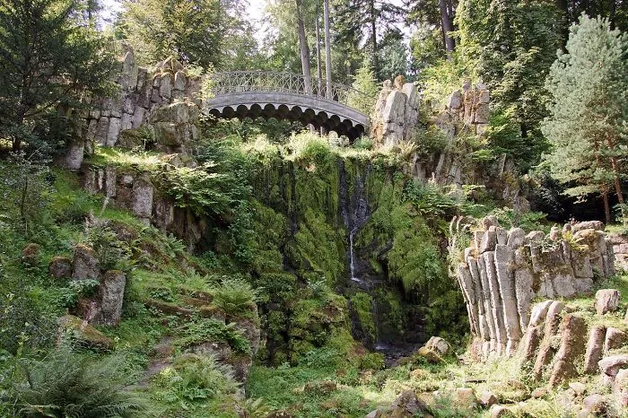 Stone mystical bridge surrounded by lush greenery and moss-covered rocks in a forest setting with a small waterfall below.