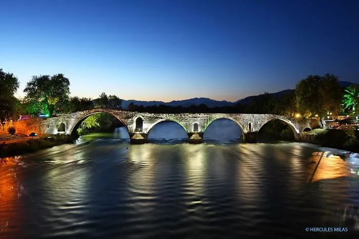 Ancient mystical bridge illuminated at twilight over calm river with mountains in the background.