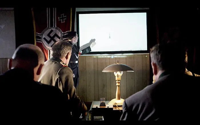 Nazi officers in a dimly lit room, one pointing at a map on the wall.
