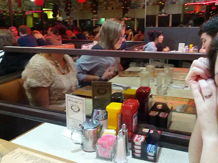 Restaurant table with unusual mirror reflection, condiment bottles, menus, and seated diners create an unexpected optical illusion.