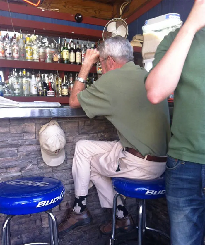 Man sitting unusually on a bar stool wearing mismatched socks, with a hat on the counter next to him.