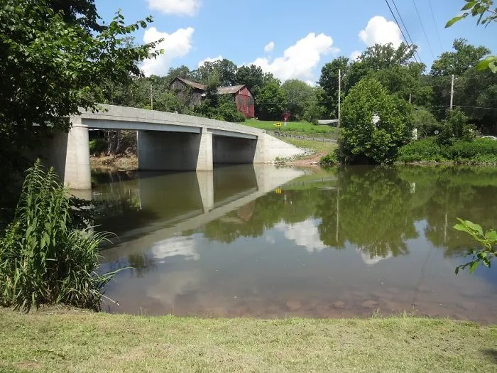 Concrete bridge over calm river surrounded by lush greenery and blue skies, a mystical bridge in a serene natural setting.