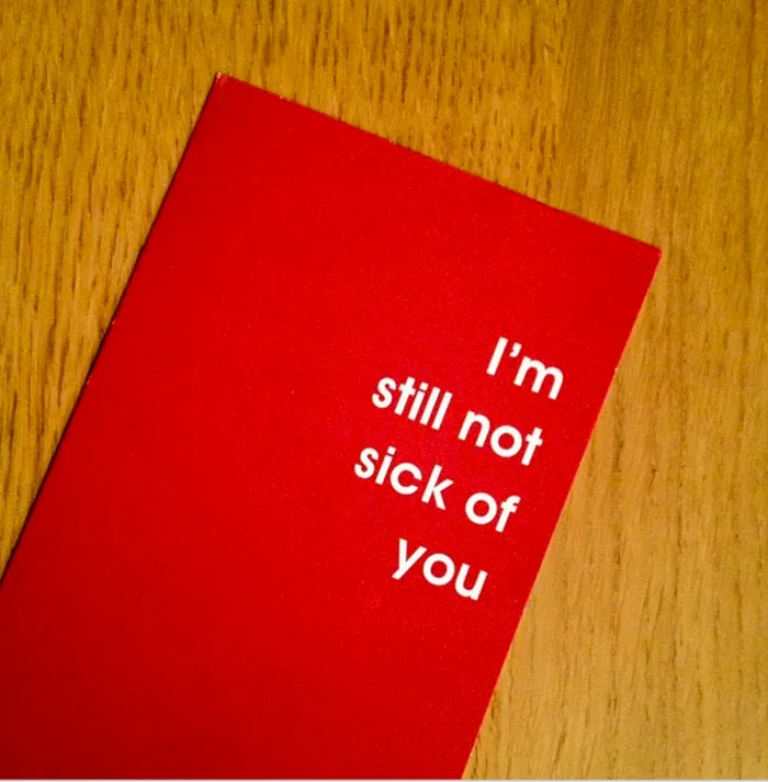 Red Valentine's Day card with white text saying "I'm still not sick of you," lying on a wooden surface.