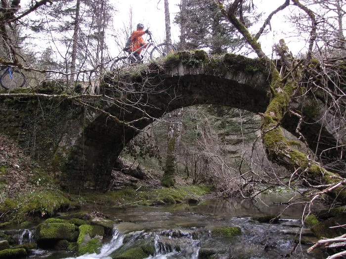 Ancient mystical bridge covered in moss over a flowing stream with a cyclist crossing in a forest setting.