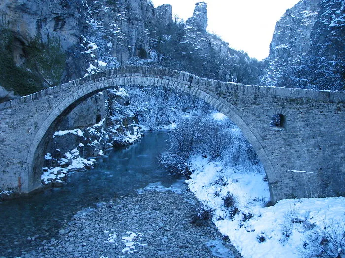 Stone arch bridge covered in snow over a serene winter river surrounded by mystical rocky cliffs and trees.